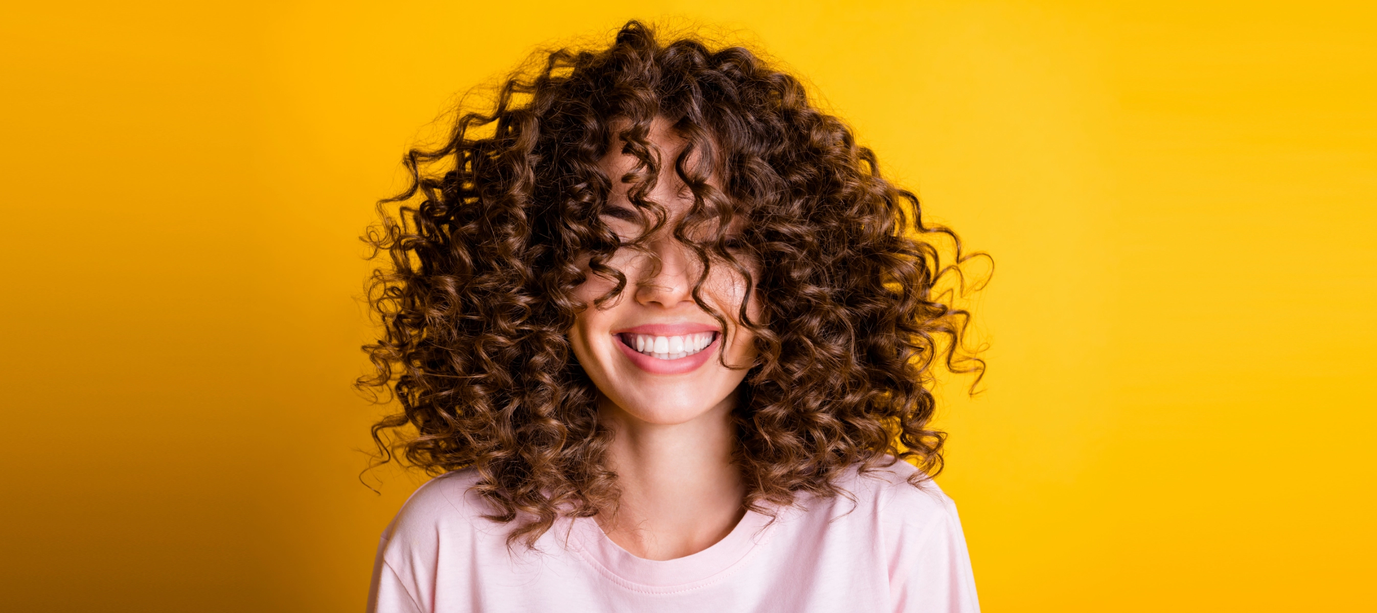 woman with 3a curly hair smiling with her eyes closed as she stands in front of a yellow background 